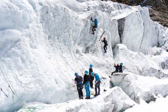 Mehrere Personen klettern an einer steilen Eiswand, einige sichern am Boden mit Helmen und Kletterausrüstung
