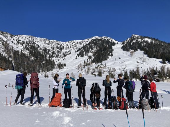Gruppe von Menschen mit Schneeschuhen und Rucksäcken steht auf verschneitem Feld vor schneebedeckten Bergen unter klarem blauem Himmel
