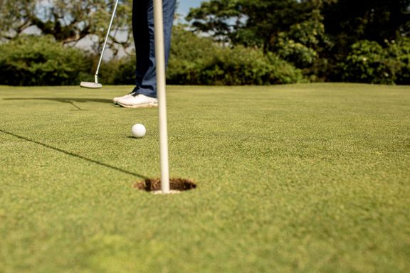 Ausschnitt eines Golfplatzes, man sieht ein Hole mit einer Stange drin und einen darauf zurollenden Golfball. Im Hintergrund sieht man die Beine und den Schläger der golfspielenden Person.
