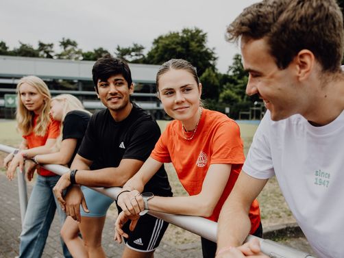 Fünf Studierende lehnen an einem Geländer im Außenbereich des Campus.
