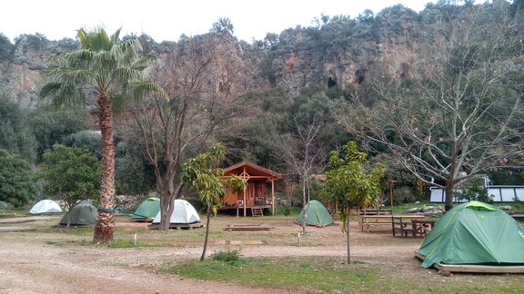 Zeltlager mit mehreren Zelten, einem Holzpavillon, Palmen und Bäumen vor einer felsigen Klippe
