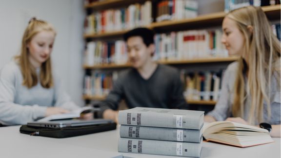 Drei Bücher liegen aufeinandergestapelt vor drei Studierenden in der Bibliothek
