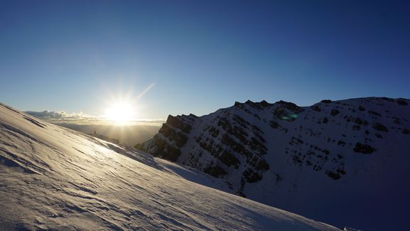Sonnenaufgang über schneebedecktem Berghang und gezackten Berggipfeln unter klarem Himmel

