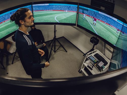 Foto von Mann der in der Mitte von sechs kreisförmig aufgestellten Monitoren mit laufender Fußballsimulation blickt
