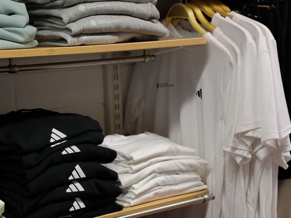 ausschnitt eines regals mit einem stapel weißen t-shirts, einem stapel schwarzen shirts mit weißem adidas-logo, einem stapel hellgrauen shirts mit weißem-spohologo; daneben hängen auf bügeln 5 weiße t-shorts mit spoho- und adidas-logo auf der brust
