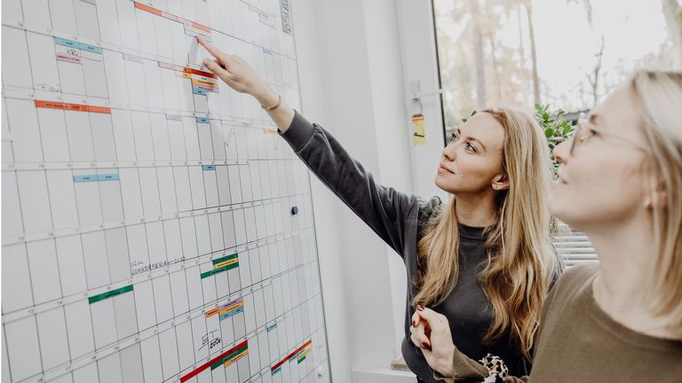 Foto von zwei Frauen die auf Terminkalender an Whiteboard zeigen
