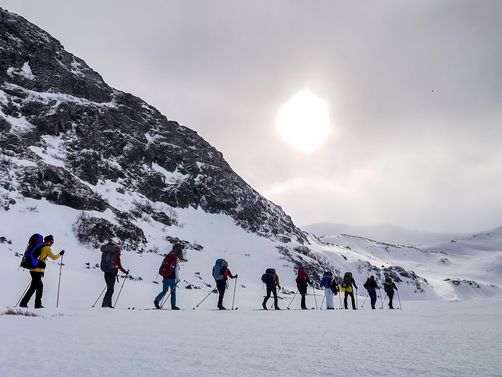 Gruppe von Wanderern mit Rucksäcken und Wanderstöcken auf schneebedecktem Gelände neben einem steilen Berg unter bewölktem Himmel
