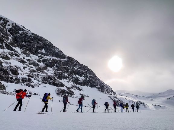 Gruppe von Wanderern mit Rucksäcken und Wanderstöcken auf schneebedecktem Gelände neben einem steilen Berg unter bewölktem Himmel
