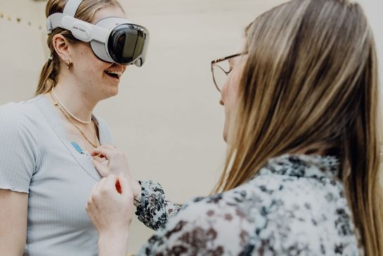 Forschende Person klebt Messelektrode an Körper einer Probandin mit VR-Brille
