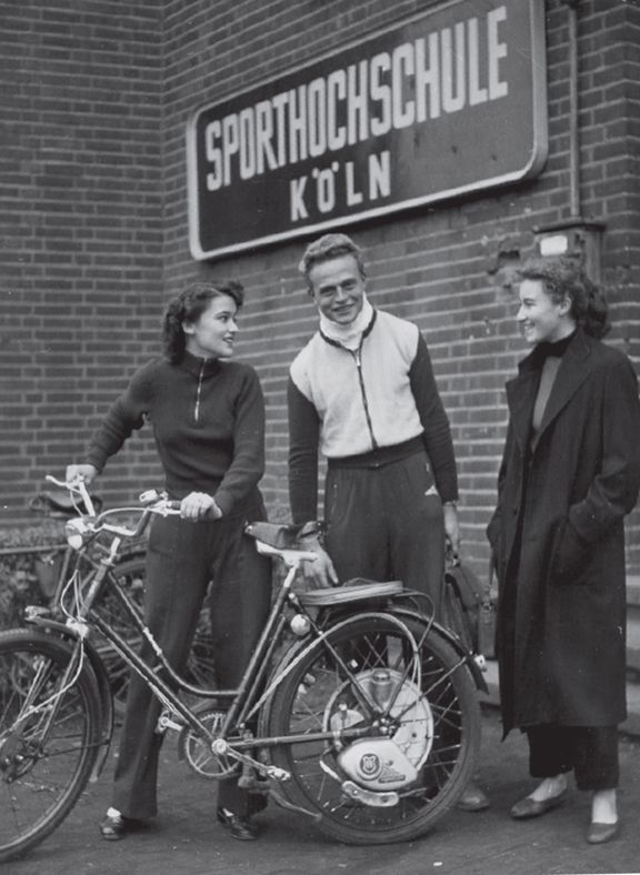 Sportstudierende mit Fahrrad circa aus dem Jahr 1960 vor Gebäude-Mauer mit Schild "Sporthochschule Köln"
