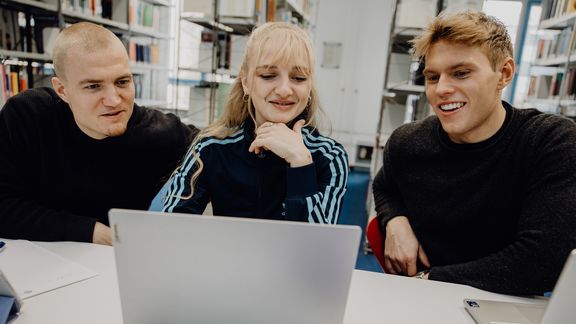 Drei Studierende sitzen vor einem Laptop in der Bibliothek der Deutschen Sporthochschule
