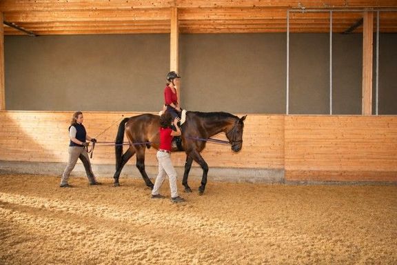 Eine Reiterin auf einem Pferd in einer Reithalle. Das Pferd wird von einer weiblichen Person, die hinter dem Pferd geht, an Leinen geführt. Eine zusätzlich weibliche Person geht neben dem Pferd und hält die Reiterin fest. 
