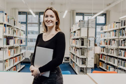 Studentin lächelt mit Laptop in der Hand in der Bibliothek
