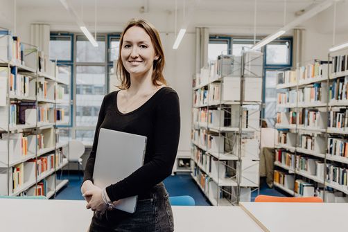 Studentin lächelt mit Laptop in der Hand in der Bibliothek
