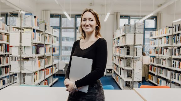 Studentin lächelt mit Laptop in der Hand in der Bibliothek
