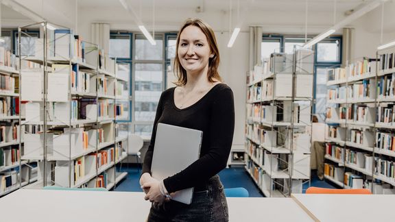 Studentin lächelt mit Laptop in der Hand in der Bibliothek
