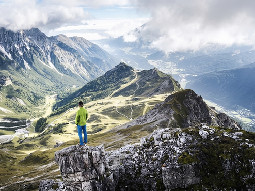 Person in grüner Jacke steht auf Felsen und blickt auf alpine Berglandschaft mit Tälern und Gipfeln
