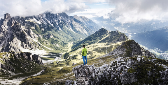 Person in grüner Jacke steht auf Felsen und blickt auf alpine Berglandschaft mit Tälern und Gipfeln
