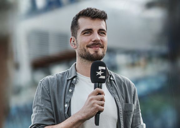Porträtfoto von Markus Paszehr. Er trägt ein weißes T-Shirt mit einer grauen Jacke darüber und hat ein schwarzes Mikrofon in der Hand.
