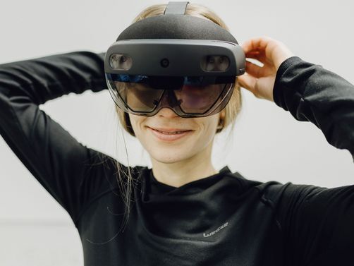 Foto von Frau die lächelnd mit großer VR-Brille in Kamera blickt 
