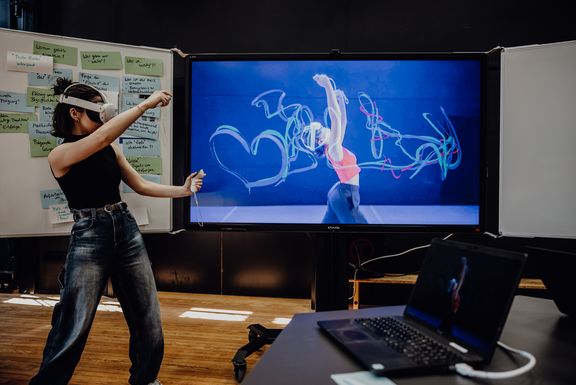 Eine Frau mit VR-Brille und Controller steht vor einem großen Bildschirm, auf dem eine tanzende Person mit digitalen Lichtspuren zu sehen ist – im Hintergrund ein Whiteboard mit Moderationskarten.
