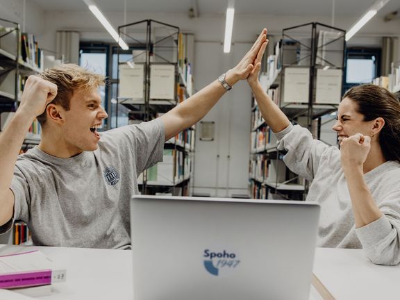 Zwei Studierende geben sich lächelnd in der Bibliothek ein High-Five

