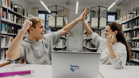 Zwei Studierende geben sich lächelnd in der Bibliothek ein High-Five
