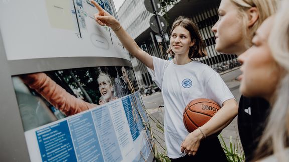 Mehrere Studierende stehen vor einer Informationstafel im Außenbereich und zeigen auf Inhalte.
