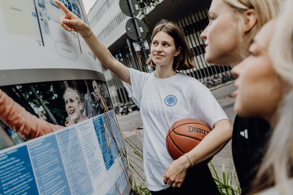 Mehrere Studierende stehen vor einer Informationstafel im Außenbereich und zeigen auf Inhalte.
