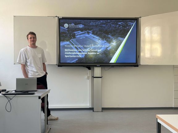 Konstantin Rehlinghaus vor einem Smartboard mit der Titelfolie seiner Präsentation
