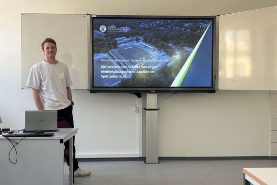 Konstantin Rehlinghaus vor einem Smartboard mit der Titelfolie seiner Präsentation
