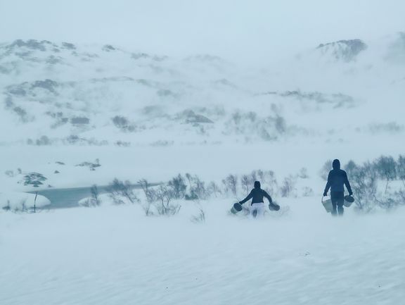 Zwei Personen in Winterkleidung tragen Eimer durch eine verschneite Landschaft mit Hügeln im Hintergrund
