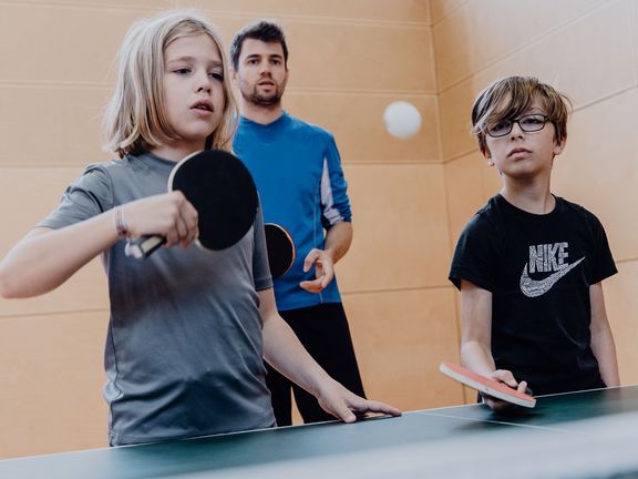 Zwei Schüler stehen vor einer Tischtennisplatte mit Schlägern und versuchen einen Ball zu treffen während der Lehrer dahinter zuschaut
