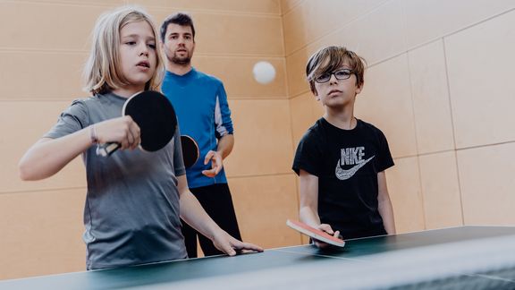 Zwei Schüler stehen vor einer Tischtennisplatte mit Schlägern und versuchen einen Ball zu treffen während der Lehrer dahinter zuschaut
