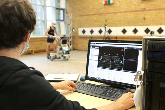 Eine Person, die vor einem Notebook sitzt und Daten analysiert, im Hintergrund eine Person mit dem Lernroboter RoSyLerNT
