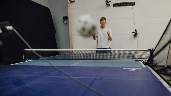 Proband führt Tischtennisaufschlag auf einer Platte in Richtung eines Netzes auf der anderen Seite aus
