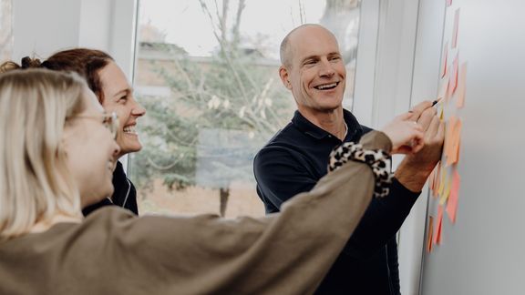 Foto von drei lächelnden Personen die auf keine Zettel an der Wand zeigen
