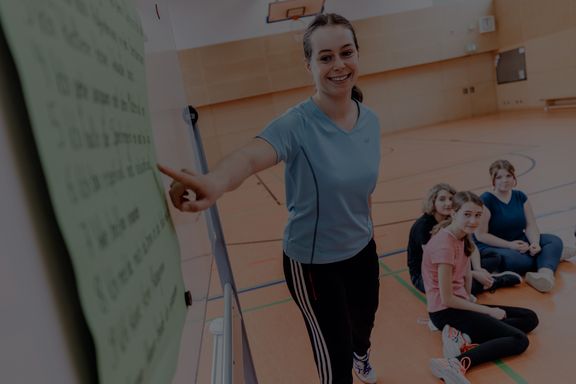 lehrerin steht in einer sporthalle an der tafel und schüler*innen sitzen interessiert auf dem boden
