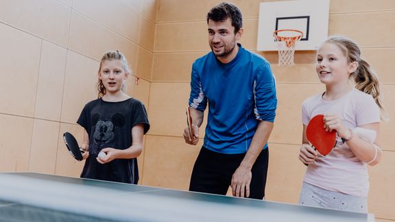 Ein Lehrer und zwei Schülerinnen stehen mit Tischtennisschlägern vor einer Tischtennisplatte
