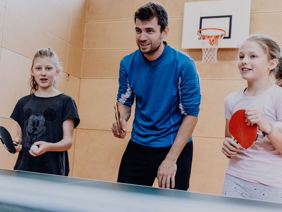 Ein Lehrer und zwei Schülerinnen stehen mit Tischtennisschlägern vor einer Tischtennisplatte

