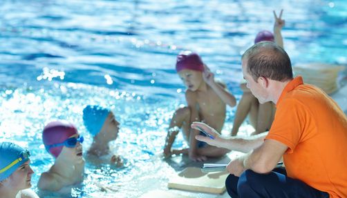 Lehrsituation im Schwimmbad: 5 Schüler*innen mit Badekappe und 1 Schwimmlehrer
