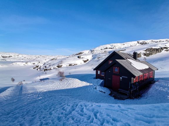 Schwarze Hütte mit roten Fensterrahmen steht im verschneiten, hügeligen Gelände unter blauem Himmel
