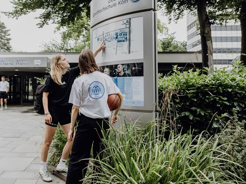 Zwei Studierende zeigen auf den Wegweiser im Eingangsbereich der Deutschen Sporthochschule
