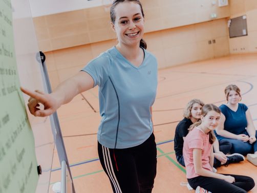 Lehrerin zeigt vor Schülern und Schülerinnen auf Plaktat in einer Sporthalle  
