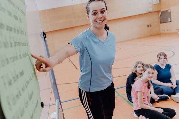 Lehrerin zeigt vor Schülern und Schülerinnen auf Plaktat in einer Sporthalle  
