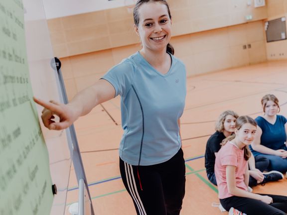 Lehrerin zeigt vor Schülern und Schülerinnen auf Plaktat in einer Sporthalle  
