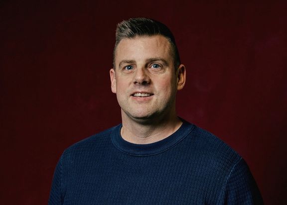 Porträtfoto von Sebastian Kleemann. Er trägt einen blauen Pullover und steht vor einem dunkelroten Hintergrund.
