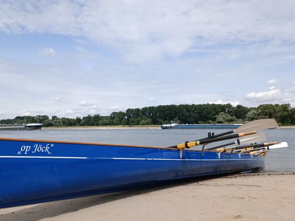 Blaues Ruderboot mit dem Schriftzug 'op Jöck' liegt am sandigen Ufer eines breiten Flusses. Mehrere Ruder sind auf der rechten Seite des Bootes befestigt. Im Hintergrund verläuft eine Baumreihe entlang des gegenüberliegenden Ufers, auf dem Wasser fahren zwei Frachtschiffe. Der Himmel ist leicht bewölkt.
