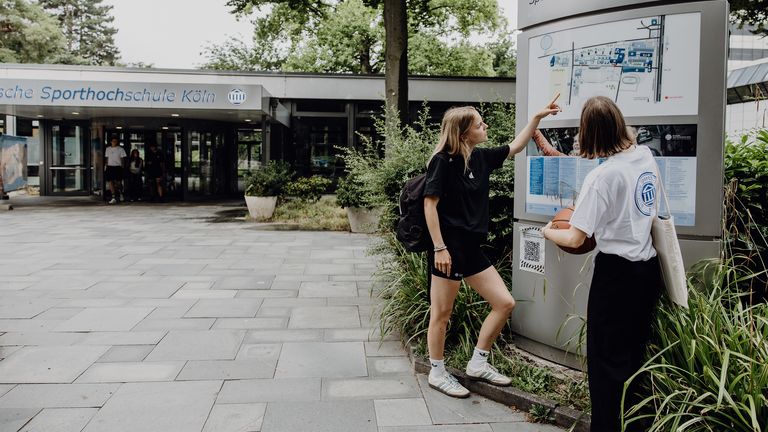 Zwei Studierende zeigen auf den Wegweiser im Eingangsbereich der Deutschen Sporthochschule
