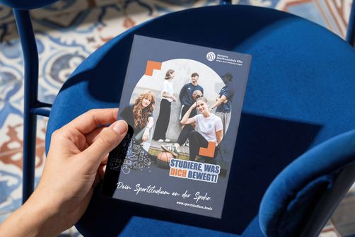 mit linker hand wird eine broschüre über einen blauen stuhl gehalten, der auf einem boden mit bunten fliesen steht
